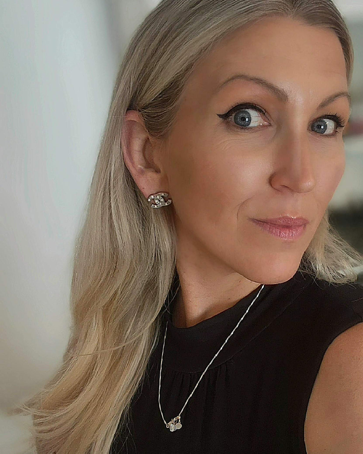 A close-up portrait of a blonde woman with blue eyes, wearing a black sleeveless top, a silver necklace, and a small silver stud earring, set against a softly blurred indoor background.